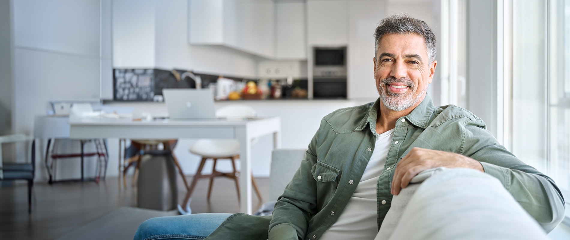 The image features a man seated on a couch with his legs crossed, wearing a denim jacket over a green shirt, smiling at the camera with his arms resting on his knees.