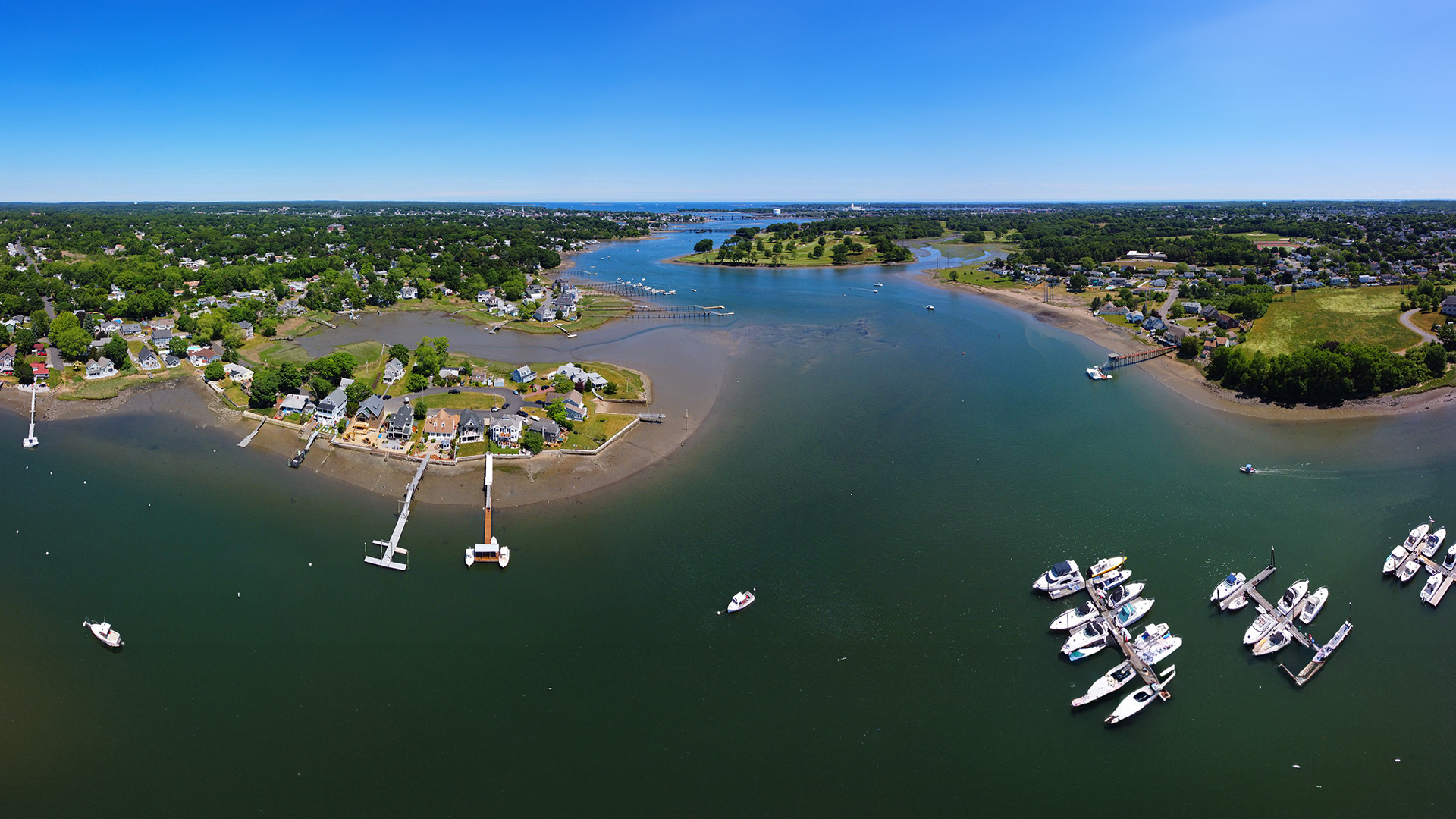 The image shows a scenic view of a river with boats docked at a marina, surrounded by houses and set against a clear sky during daytime.