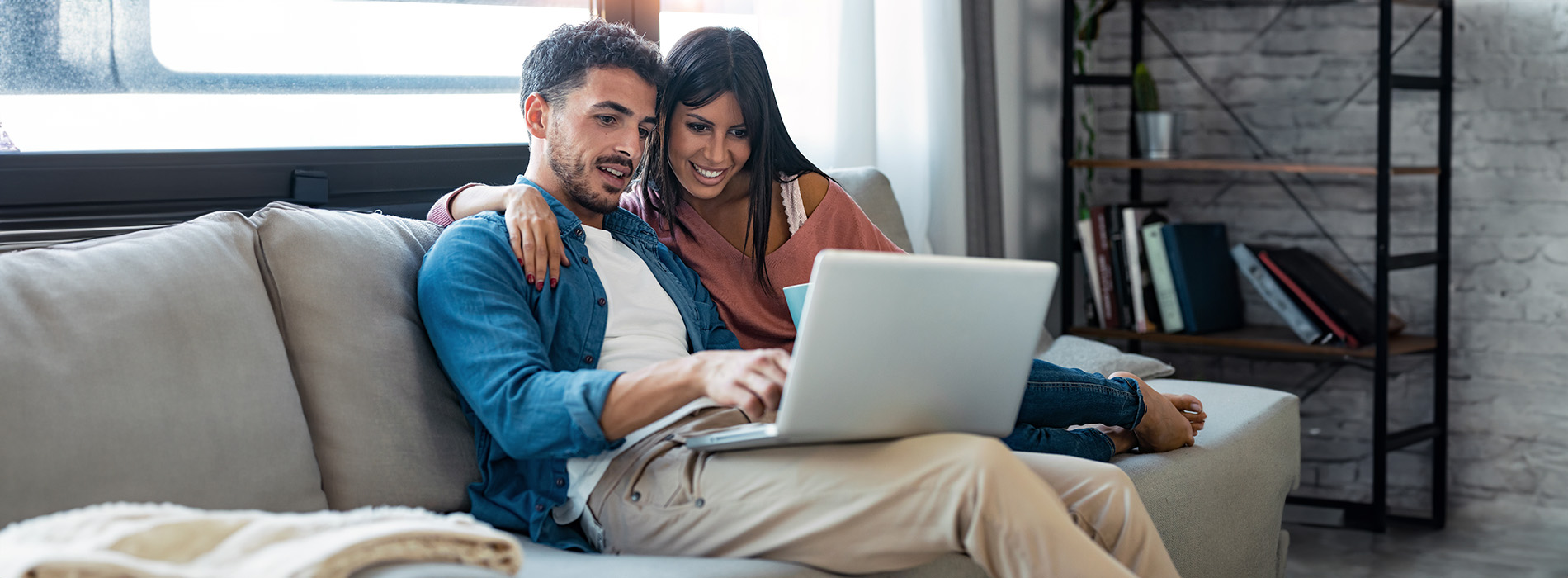 The image depicts a man and woman sitting on a couch with a laptop between them, both smiling and looking at the camera.