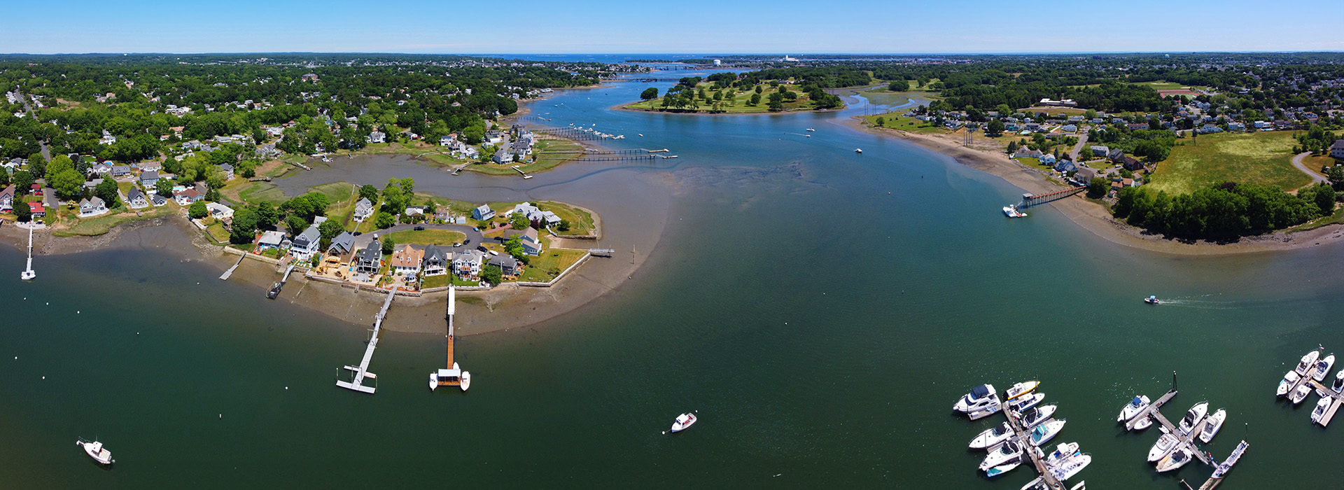 The image depicts a scenic waterfront view with boats docked at a pier, surrounded by houses on the shoreline, under a clear sky.
