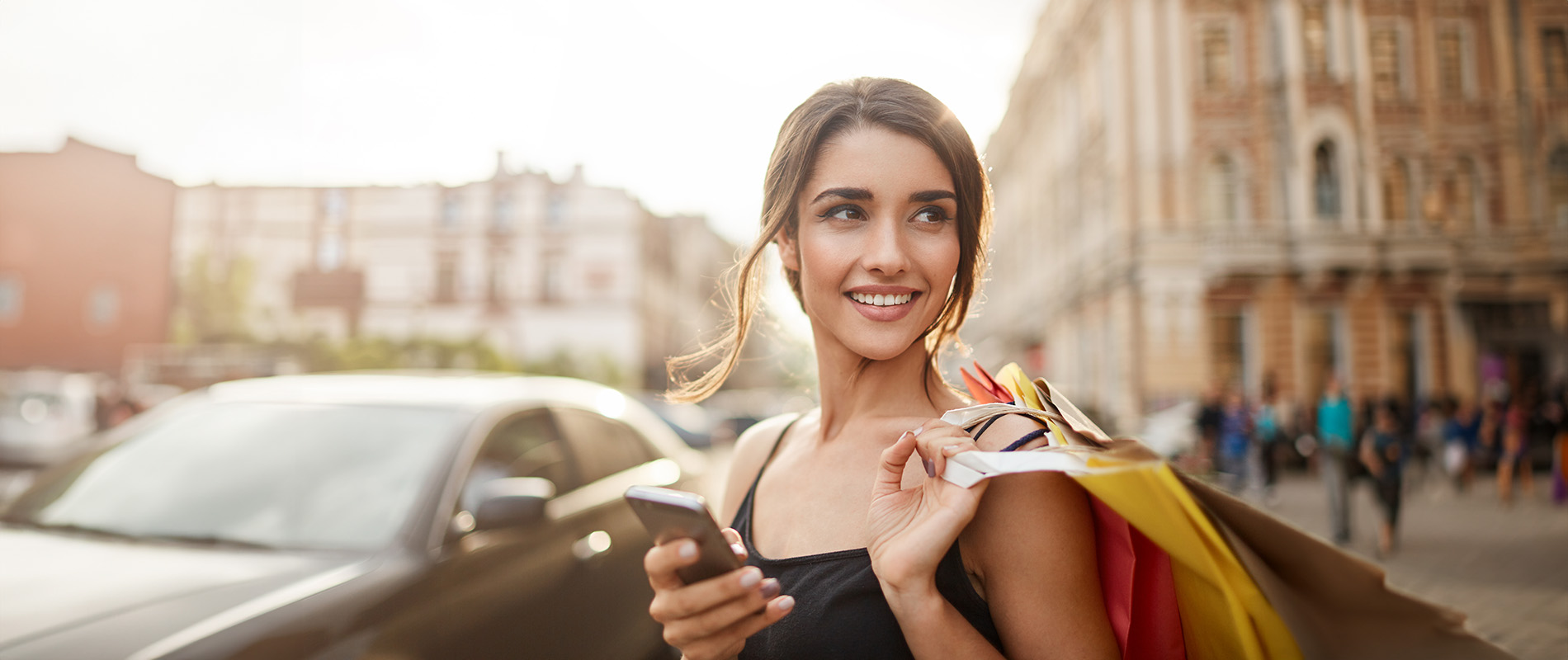 The image shows a woman walking down a street, smiling at the camera, with her hand on her hip, holding a smartphone, dressed in casual attire, with a shopping bag over her shoulder, during daylight with clear skies.