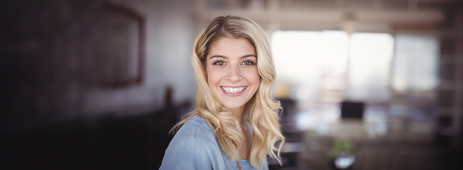 A smiling woman with blonde hair stands in an indoor setting, looking directly at the camera.
