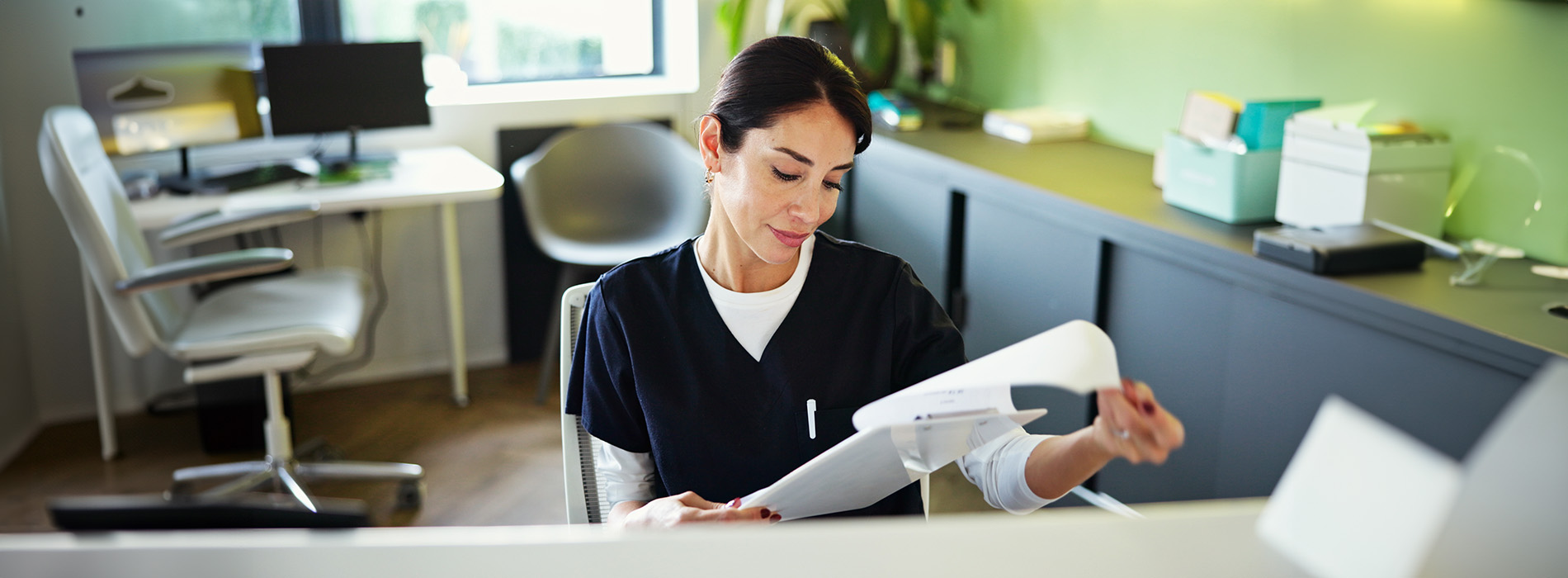 The image shows a woman sitting at an office desk, looking at papers, with her hands on a clipboard, set against an interior office environment.