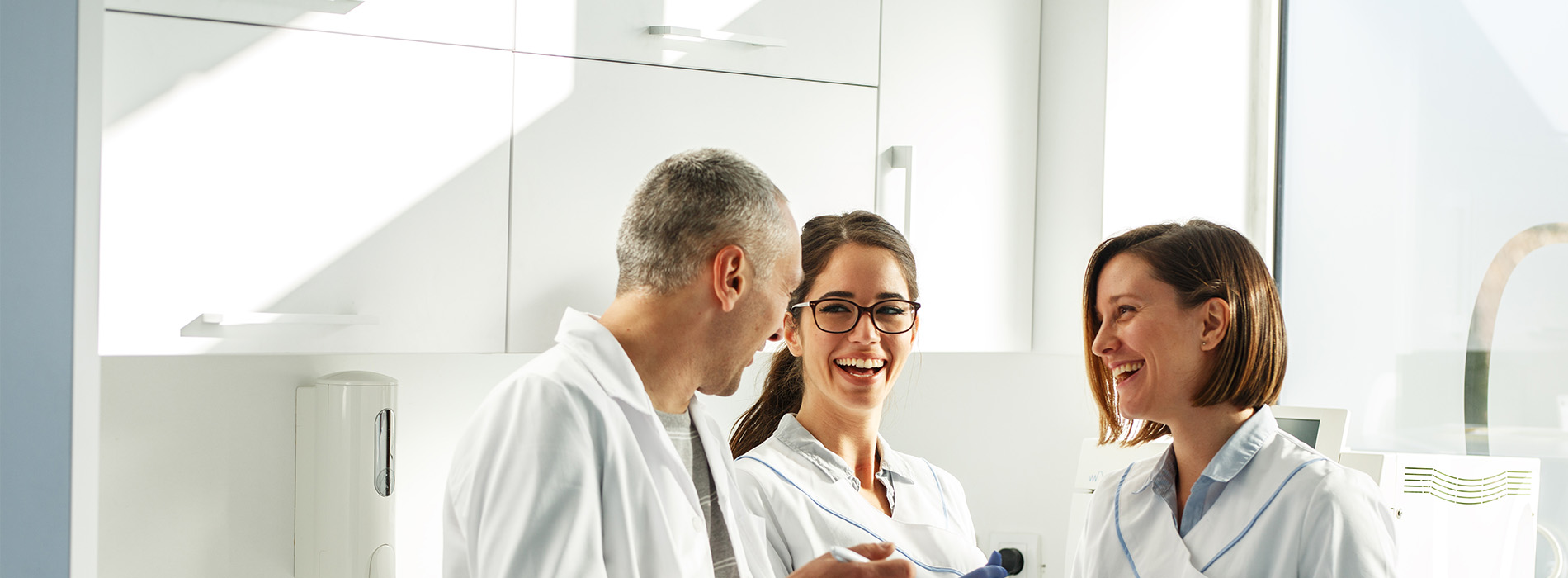 The image shows four individuals, likely professionals, engaged in conversation inside a brightly lit room with white walls, which appears to be an office setting.