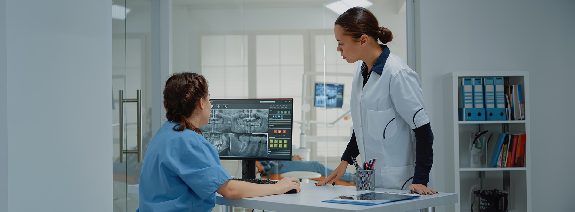 The image depicts three individuals in a medical setting with screens displaying information, likely a hospital environment where they are engaged in work-related activities.