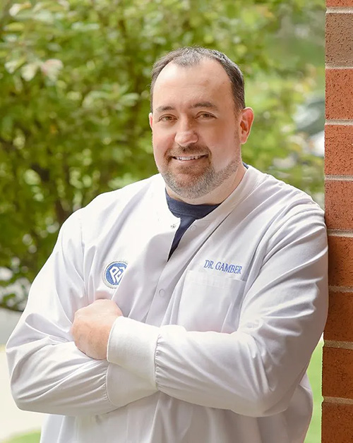 The image shows a man wearing a white lab coat standing outdoors with his arms crossed in front of him.