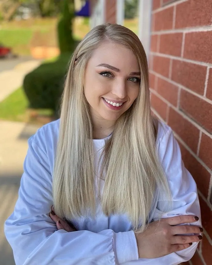 A young woman with blonde hair poses confidently against a brick wall, smiling at the camera.