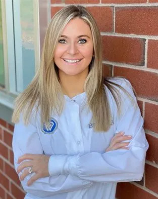 A smiling woman wearing a white lab coat poses confidently with her arms crossed against a brick wall.