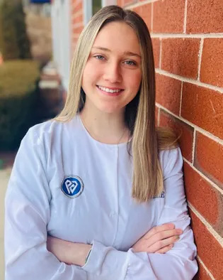 The image shows a woman standing against a brick wall with her arms crossed, wearing a white lab coat with a logo on the left chest area, smiling at the camera.