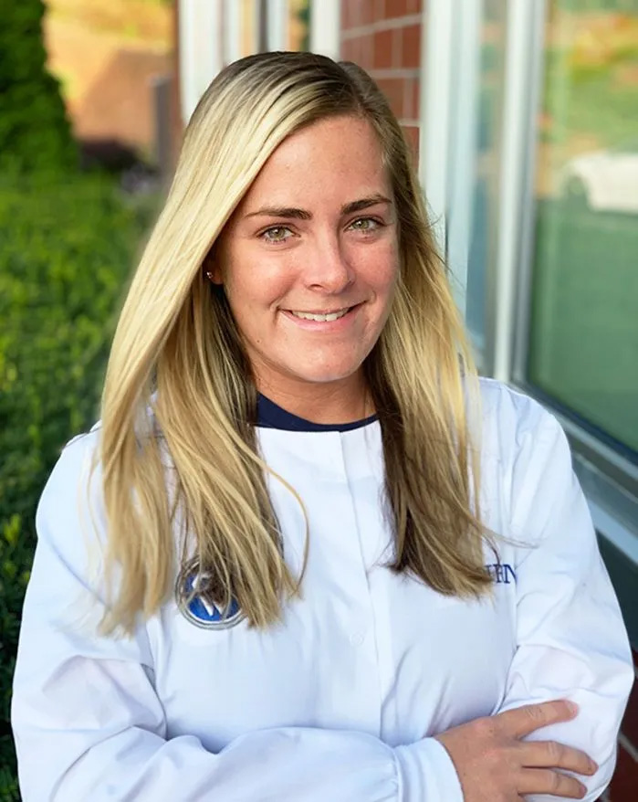 A woman wearing a white jacket with a logo on the left chest area, smiling at the camera while standing outdoors against a brick wall.