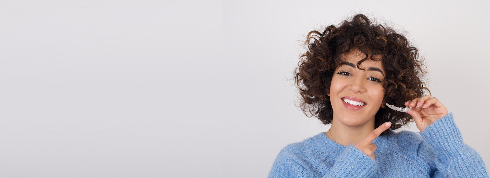 This is a photographic image of a person with curly hair smiling at the camera while holding a toothbrush, wearing a blue top, against a plain white background.
