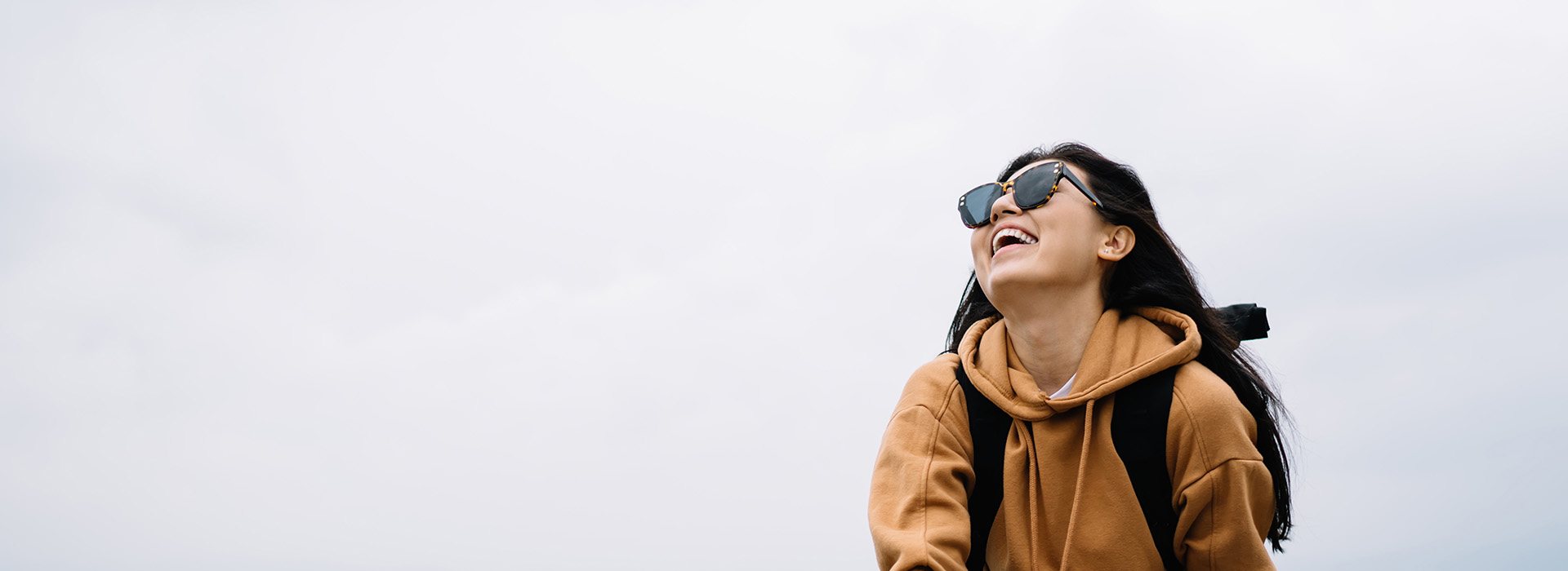 A person wearing sunglasses and a backpack, smiling while looking upwards, against a sky background with clouds.