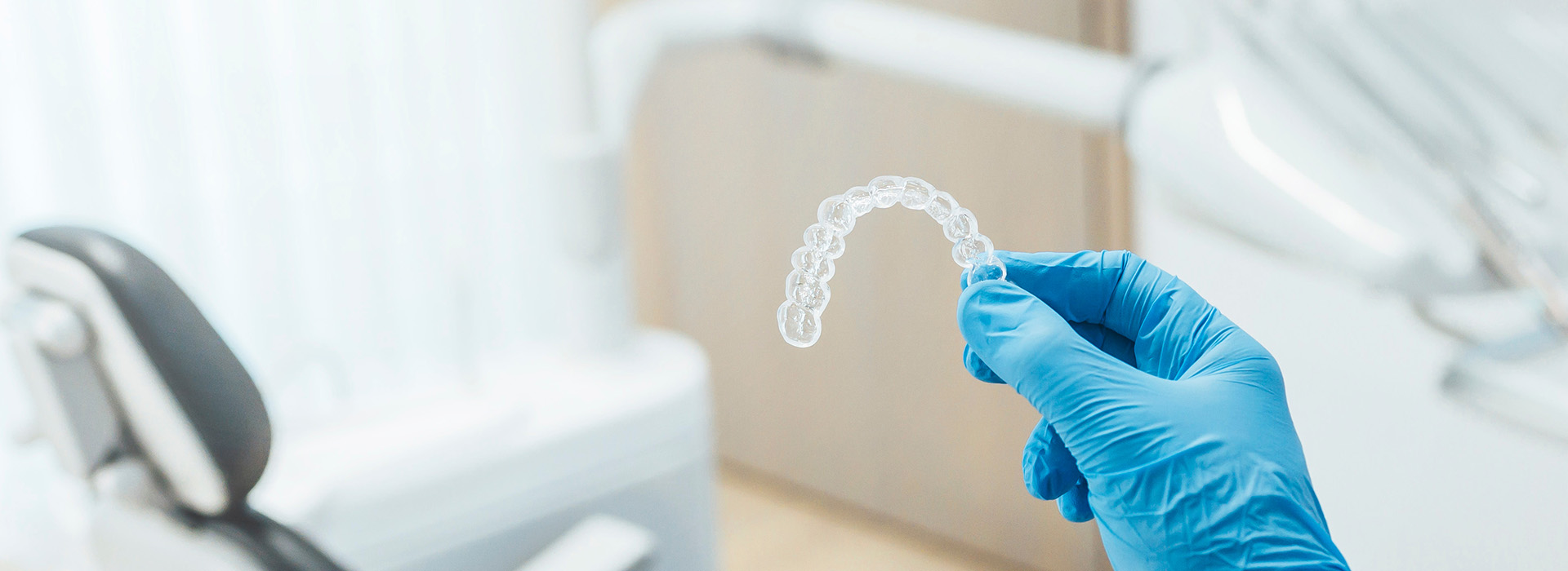 The image shows a hand wearing blue gloves holding a transparent object with a white substance inside, which appears to be a dental implant, against a blurred background of a medical or dental setting.
