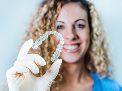 The image shows a woman holding up a clear dental retainer with her hand, smiling at the camera.