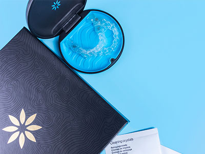 The image shows a blue box with a black lid containing a small item resembling a jar of crystals, placed on a light blue surface next to a clear plastic container with a blue lid that has a green substance inside, and there s a blue object that appears to be a decorative item or an accessory in the background.