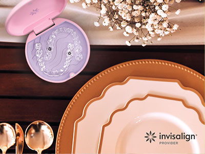 This is a photograph featuring an assortment of tableware items including plates, bowls, and a cake stand with a decorative lid, displayed on a wooden surface with a pinkish hue. The background shows a portion of a dining table and some decorative elements like flowers and a vase.