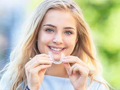 The image shows a young woman with a radiant smile, holding up a toothbrush with a smiley face on it.