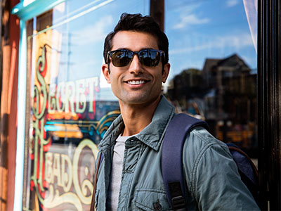 The image shows a young man standing outdoors in front of a shop window, wearing sunglasses and holding his backpack. He has a beard and mustache, is smiling, and appears to be posing for the photograph.