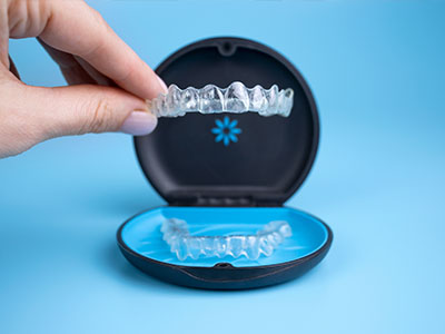 The image shows a person s hand holding an open box containing a clear plastic dental retainer with teeth impressions, placed on a blue background.
