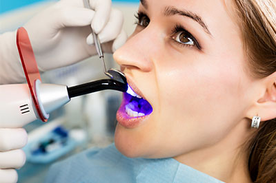The image shows a person receiving dental treatment, with a dentist using a device to clean or examine their teeth while wearing gloves and a face mask.