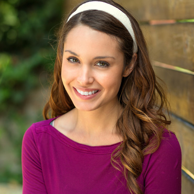 A smiling woman with long brown hair wearing a purple top and a white headband poses for the camera.