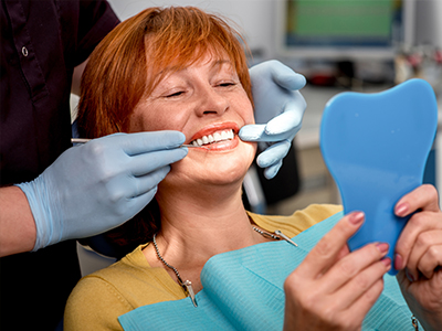 The image shows a woman seated in a dental chair with a smile on her face, holding up a blue object towards the camera. She appears to be engaged in a dental appointment, as indicated by the presence of dental tools and personnel around her.