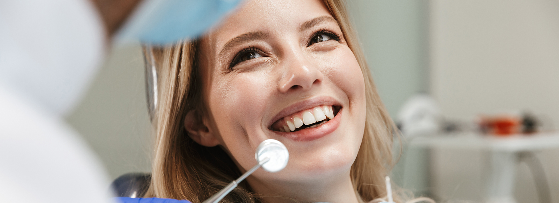 A woman is seated in a dental chair with a smile on her face, looking at the camera while a dentist examines her teeth.