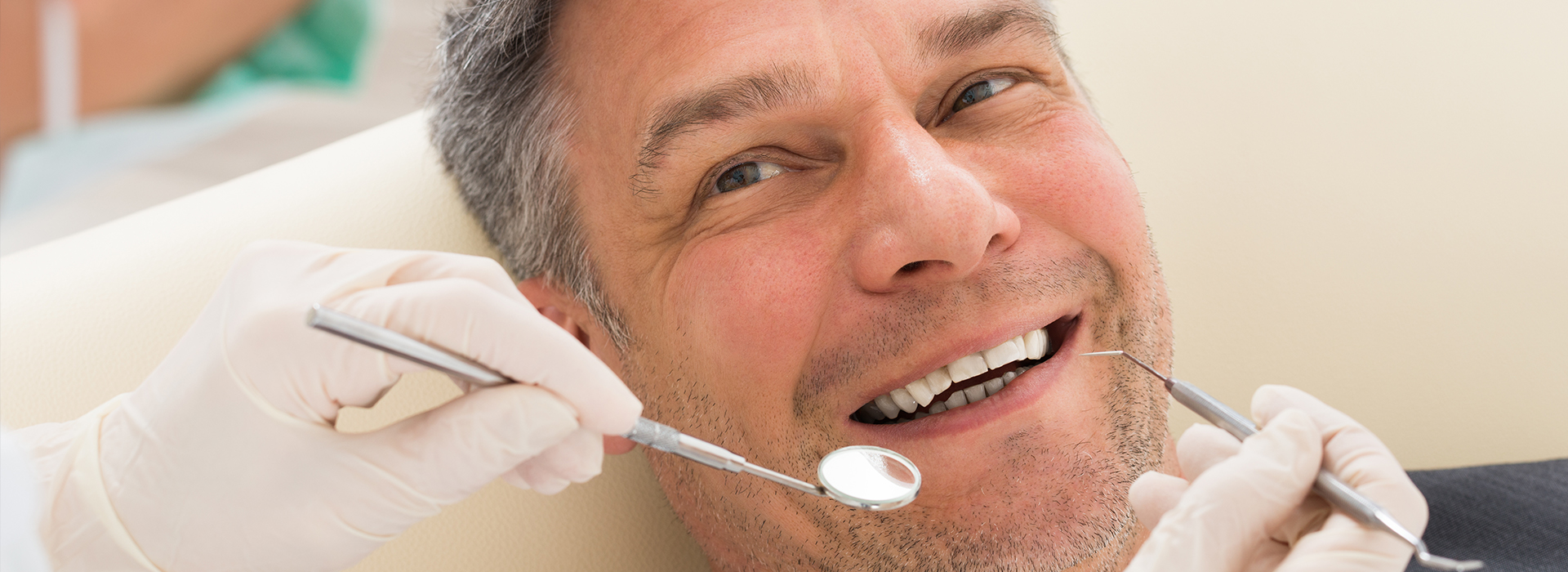 A man receiving dental care, seated in a dental chair with a dentist s hands visible, smiling while looking at the camera.