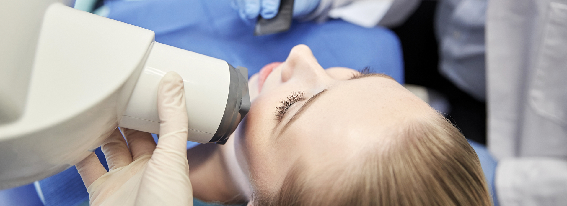 A person receiving a facial treatment under a magnifying device, with medical professionals attending to them in a clinical setting.