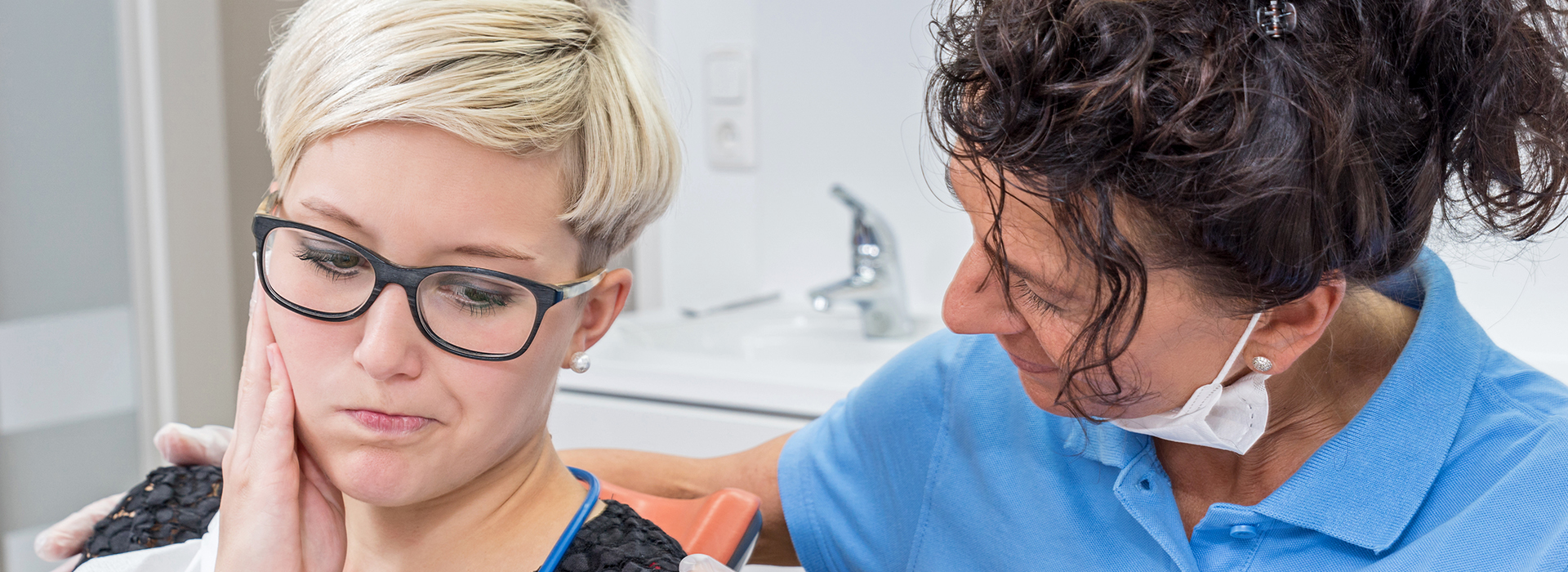 An adult woman with glasses is seated in front of a dental professional, who appears to be conducting an examination on her teeth.