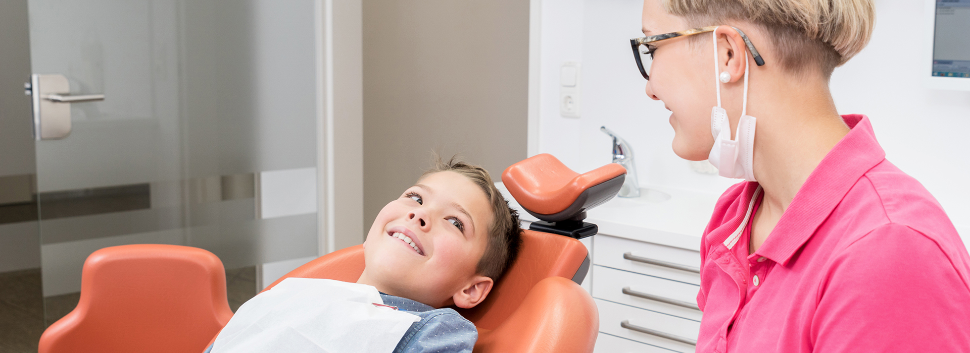 The image shows three individuals  a woman seated in a dental chair with her eyes closed, a child sitting beside her also in a dental chair, and a person standing behind them who appears to be a dental professional. They are all in an office setting, likely a dental clinic, as indicated by the equipment and the environment.