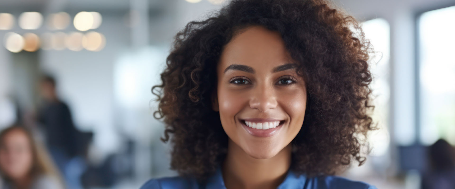 The image shows a smiling woman with curly hair, wearing a blue top, standing in an office environment with colleagues in the background.