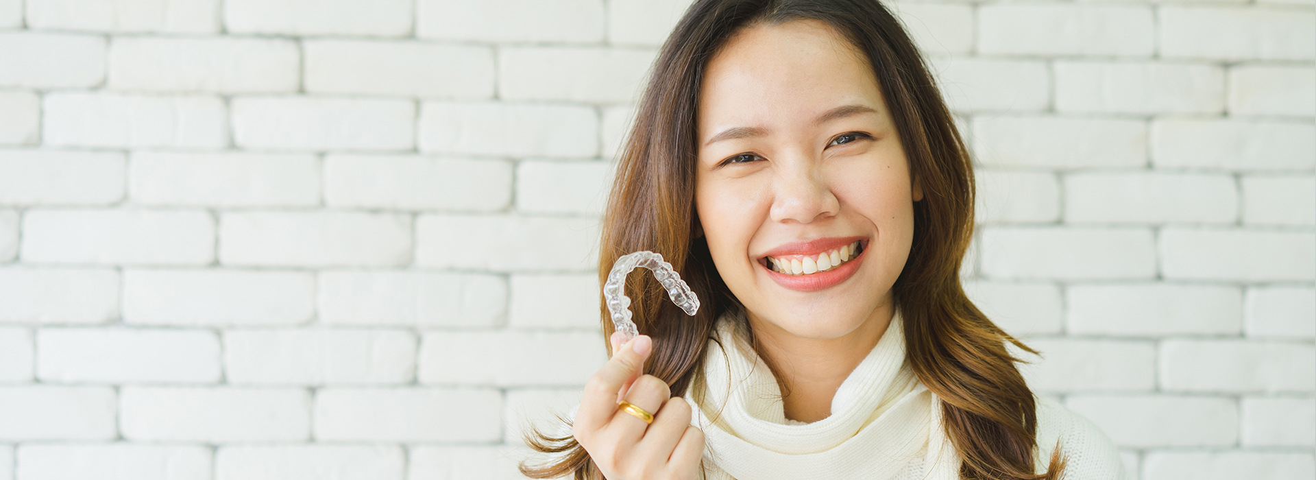 A woman with a smile holds up a toothbrush against her face, set against a white brick wall.