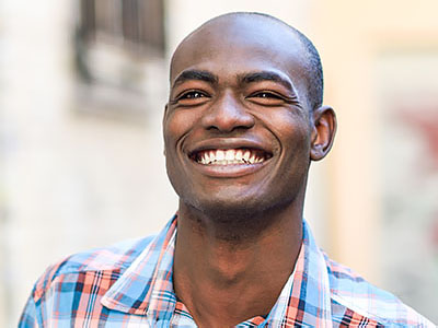 The image shows a smiling man with short hair, wearing a blue plaid shirt, standing outdoors during daylight.