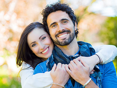 A man and woman are embracing each other outdoors, with the man wearing a blue shirt and the woman in a white top.