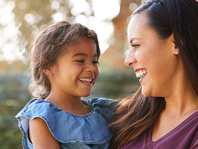 The image is a photograph featuring an adult woman and a young child, both smiling, with the woman holding the child in her arms. They appear to be outdoors during daylight, with the woman looking down at the child and the child looking up at the woman.