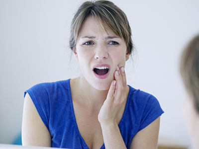 Woman with her mouth open, looking at her reflection in the mirror while holding her face with her hand.
