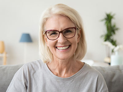 The image depicts a woman with short blonde hair, wearing glasses, smiling at the camera, set against an indoor background.
