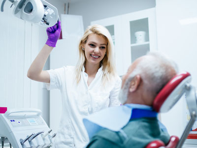 A woman in a white coat stands next to an elderly man in a dental chair, both in a clinical setting.