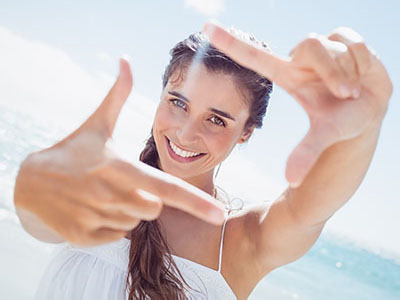 A young woman with long hair smiles while holding up her hand to create a camera viewfinder, set against a bright beach backdrop.